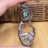 A hand holds a silver-toned wire-wrapped pendant featuring a round iridescent opal stone near the top and a larger shield-shaped stone with green, brown, and white marbled inclusions at the bottom, set against a wooden and leather background.
