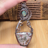 A close-up of a hand holding a silver wire-wrapped pendant featuring a large shield-shaped opal stone and a smaller round iridescent opal above it, set against a background of a wooden surface and a textured brown leather item.