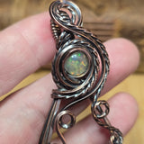 A close-up of a hand holding a handmade silver-toned pendant featuring a round cabochon opal with green and orange play-of-color, set in an ornate, twisted metal frame.