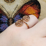 Close-up photo of a handmade double spiral copper ring worn on a finger, showing two smooth copper spirals facing each other with a polished warm metallic finish against a multicolored woven fabric background.
