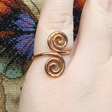 Close-up photo of a handmade double spiral copper ring worn on a finger, showing two smooth copper spirals facing each other with a polished warm metallic finish against a multicolored woven fabric background.