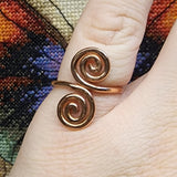 Close-up photo of a handmade double spiral copper ring worn on a finger, showing two smooth copper spirals facing each other with a polished warm metallic finish against a multicolored woven fabric background.