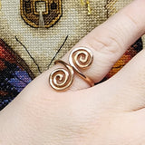 Close-up photo of a handmade double spiral copper ring worn on a finger, showing two smooth copper spirals facing each other with a polished warm metallic finish against a multicolored woven fabric background.
