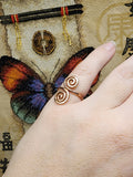 Close-up photo of a handmade double spiral copper ring worn on a finger, showing two smooth copper spirals facing each other with a polished warm metallic finish against a multicolored woven fabric background.