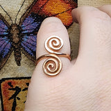 Close-up photo of a handmade double spiral copper ring worn on a finger, showing two smooth copper spirals facing each other with a polished warm metallic finish against a multicolored woven fabric background.