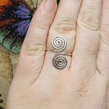 Close-up photo of a handmade double spiral sterling silver ring worn on a finger, featuring two polished silver spirals facing each other on an adjustable band, photographed against a textured multicolored fabric background.