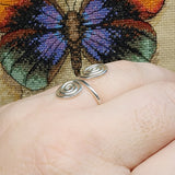 Close-up photo of a handmade double spiral sterling silver ring worn on a finger, featuring two polished silver spirals facing each other on an adjustable band, photographed against a textured multicolored fabric background.