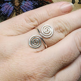 Close-up photo of a handmade double spiral sterling silver ring worn on a finger, featuring two polished silver spirals facing each other on an adjustable band, photographed against a textured multicolored fabric background.